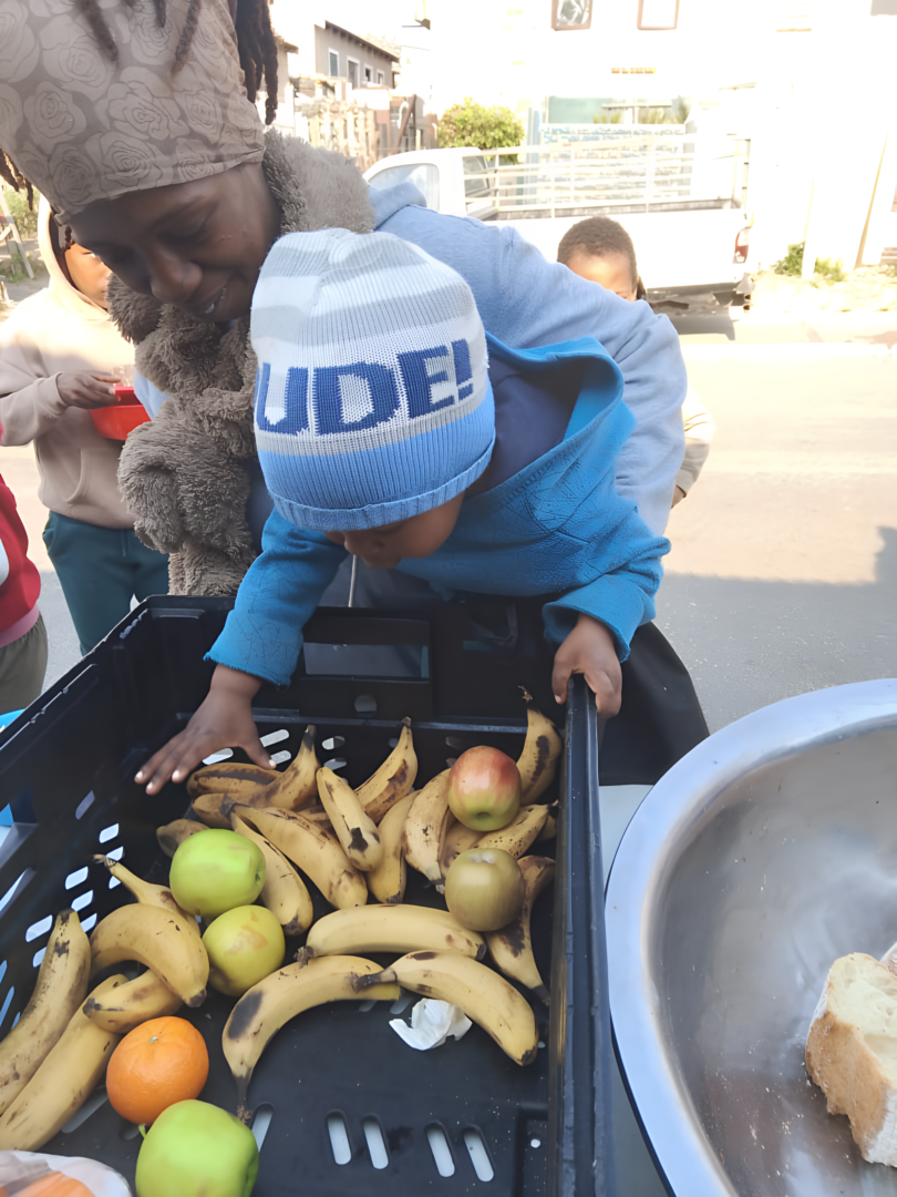 Groundbreakers – A Mother & Child Reaching into a Crate of Fruits
