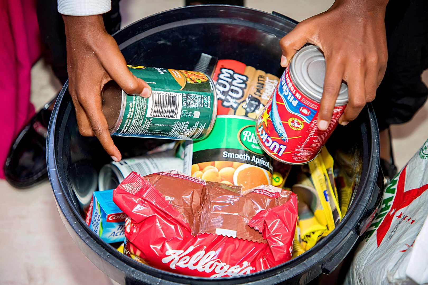 Hands placing canned food, cereal, and toiletries into a black bucket filled with essentials, representing the Buckets of Nutrition initiative supported by Estelle Ellis and SA Harvest.